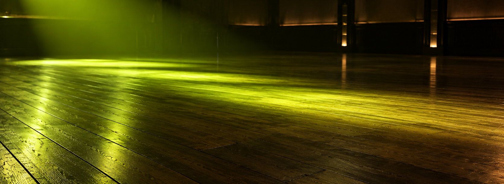 Photograph of an empty sprung dance floor seen from its edge, one green beam laid across the boards and the rest of the room in deep shadow.